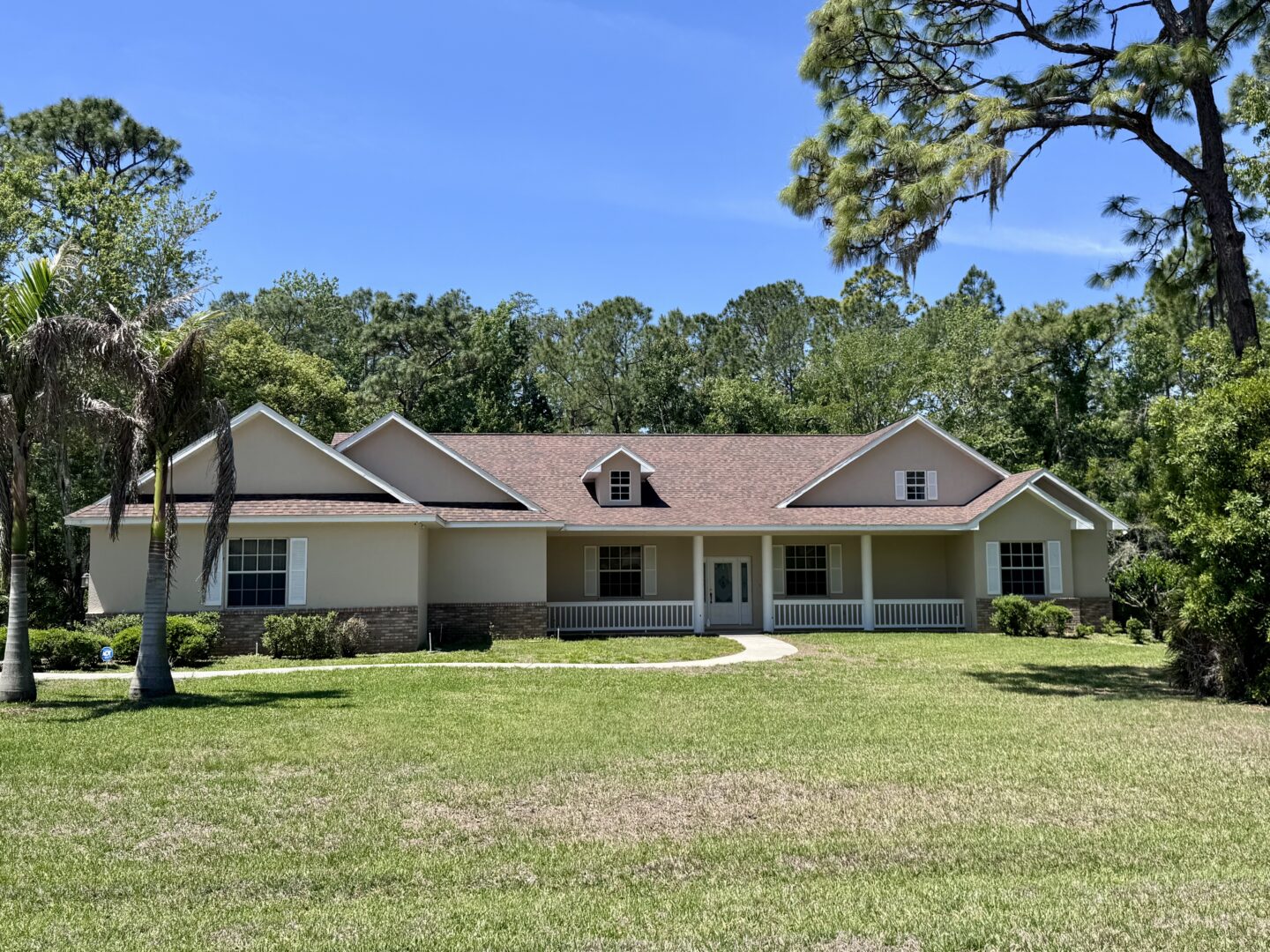 Single-story beige home with a brown shingle roof and front porch, set on a large lawn with palm trees.